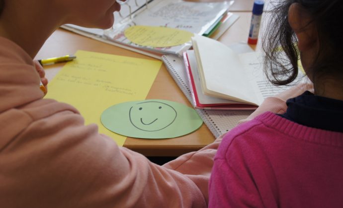 Eine Lehrkraft gibt einer Schülerin Rückmeldung an ihrem Lernplatz und nutzt dazu unter anderem eine Karte mit einem Smiley